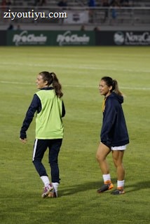 Two people are on a soccer field, wearing sports attire. One person is wearing a green vest, and the other is in a blue jacket and white shorts. The setting appears to be during a sports event, with adverts in the background and a partially filled stadium.
