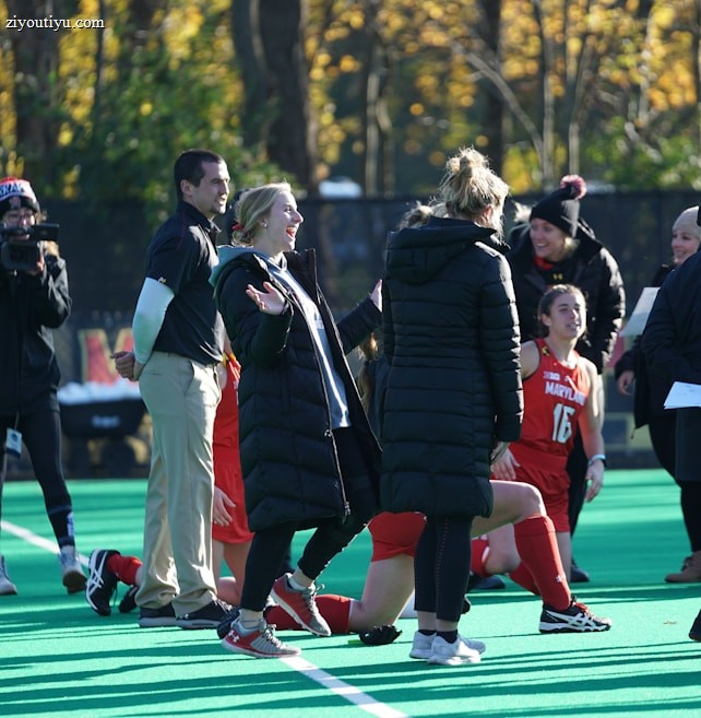 A group of people on a sports field, including several athletes in red uniforms and coaches wearing black coats. Some individuals are kneeling, while others are standing, engaged in animated conversation. A photographer or videographer is capturing the scene beside them. The background features a fence and autumn trees with yellow leaves.