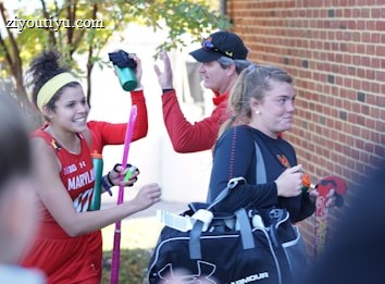 A group of athletes in sports attire appears to be in a joyful mood. One person is holding a field hockey stick and wearing a headband, while another carries a sports bag. They are gathered near a brick wall with trees in the background, sharing a light moment, likely after a sports event.