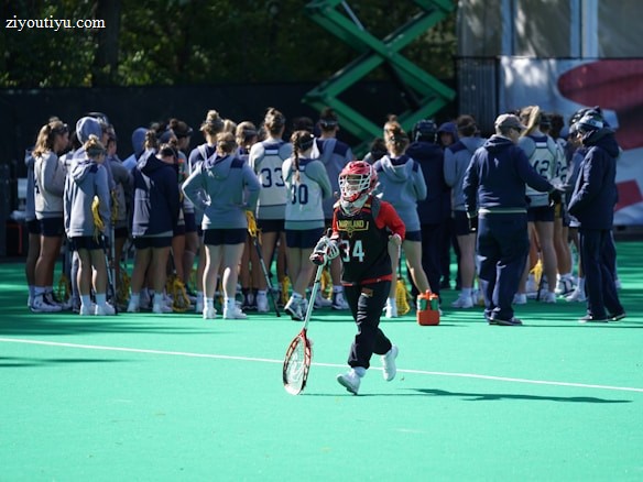 A sports team gathers on a field with a coach, all dressed in athletic gear, while one player wearing a red helmet and jersey walks away from the group carrying a lacrosse stick. The field is green, and there are sports equipment and water bottles visible.