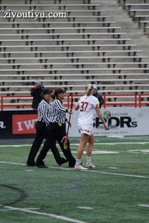 A player in a white sports uniform with the number 37 is standing on a sports field, accompanied by two referees in black and white striped shirts. The background shows empty stadium seating and advertising banners along the sidelines.