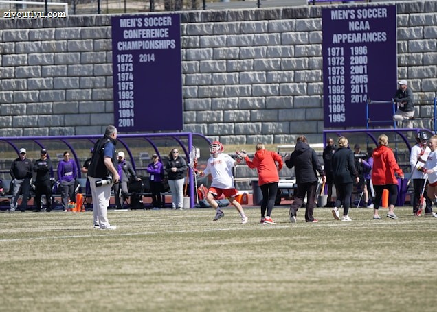 A sports team is practicing or playing on a grassy field in front of a large stone wall with banners displaying men's soccer achievements. People are wearing sports attire, and some are engaged in conversation or coaching.