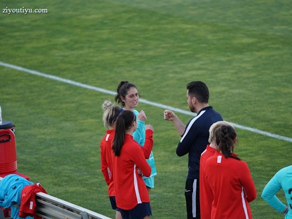 A group of people wearing sports attire are gathered on a grassy field. Some individuals are dressed in red jackets, while others are in turquoise and navy blue. A coach appears to be giving instructions. Sports equipment, such as water bottles and a bench with towels, can be seen nearby.