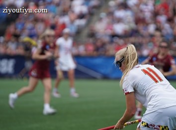 A sporting event with athletes on a grass field, one in a white jersey with the number 11 in the foreground, holding a lacrosse stick. Players in the background wear maroon uniforms. Spectators fill the stands creating a lively atmosphere.