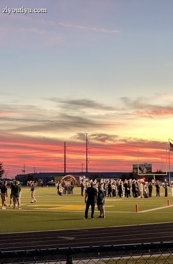 A sports field with a football game unfolding under a vibrant sunset sky, featuring a large inflatable mascot and a scoreboard displaying the score. Players and coaches are visible on the green field, surrounded by a track and some flags waving in the breeze.