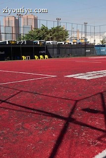 A sports court with a red playing surface and white lines, surrounded by a black mesh fence. In the background, tall buildings are visible, along with some trees and a few yellow chairs positioned near the fence.